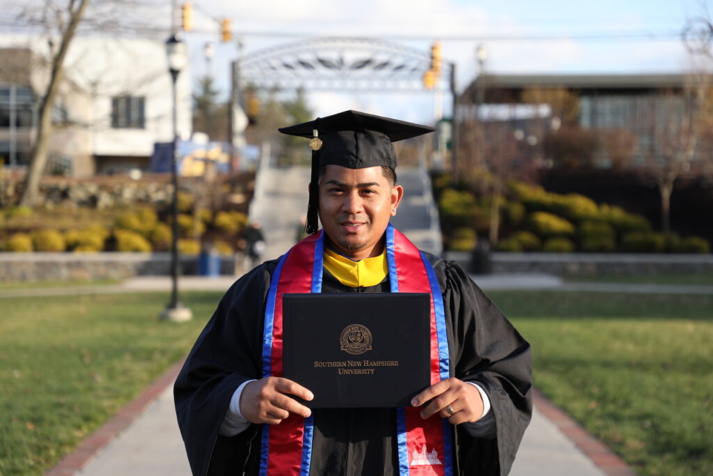 Man smiling on campus green space, wearing graduation cap and gown, holding diploma cover
