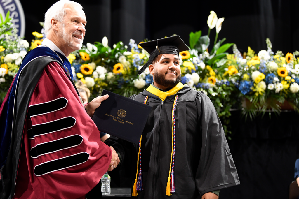 Remus Figueroa proudly accepts his diploma, shaking hands with SNHU President LeBlanc at his Commencement ceremony.
