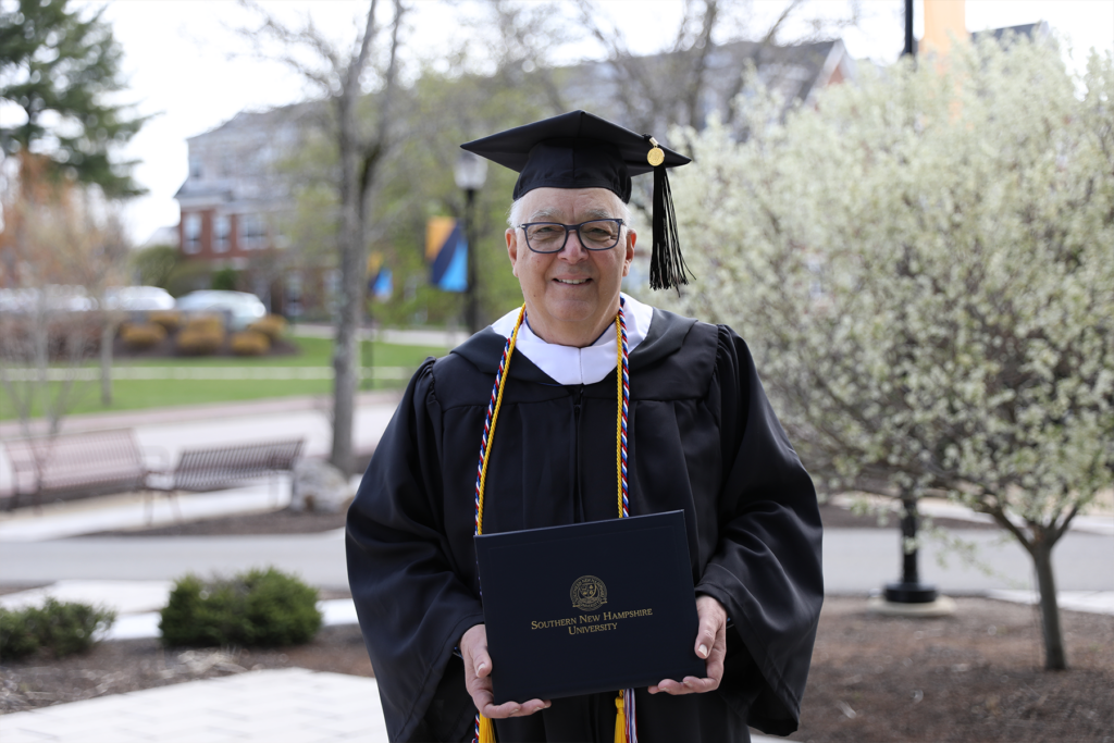Robert Heckinger, adorned in cap and gown, triumphantly holds his long-awaited diploma on the SNHU campus, radiating pride and achievement.