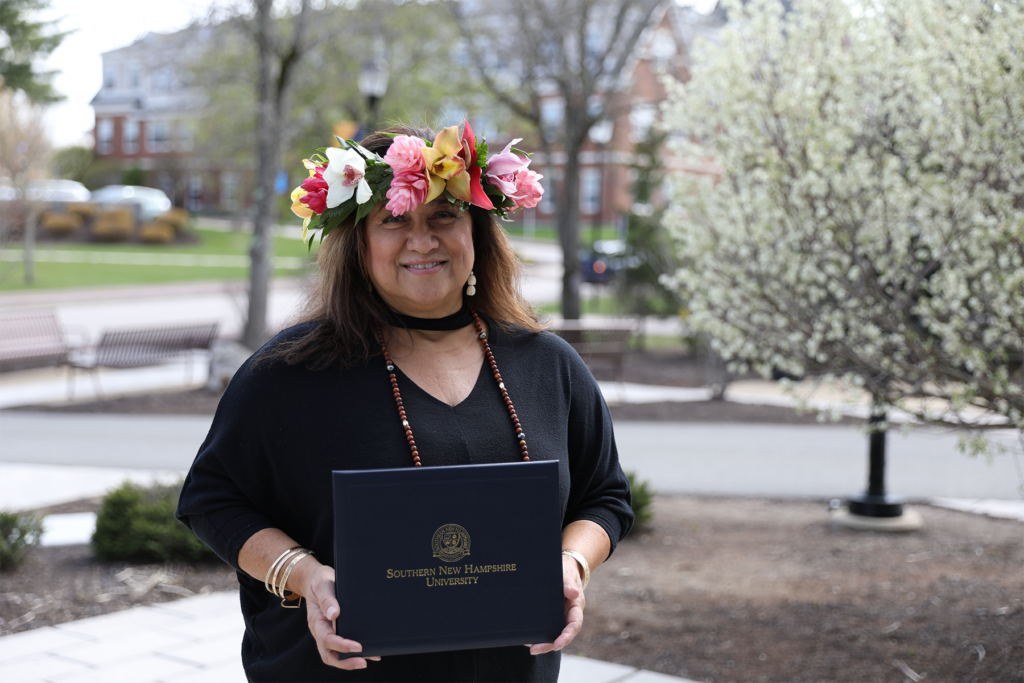 Sharla Kaleihua Kahale-Miner proudly wears a Hawaiian flower lei on her head while holding her diploma, with the SNHU campus as the backdrop.